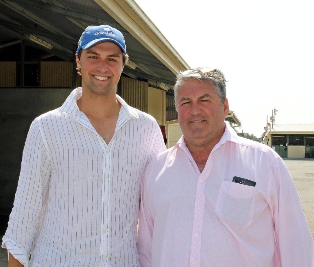 Two men stand side by side smiling outdoors one in a striped white shirt and blue cap the other in a pink shirt with a phone in his pocket