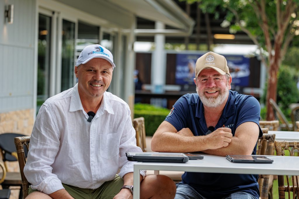 Two smiling men sit at an outdoor table with tablets in front wearing casual shirts and caps mirunners and north logos